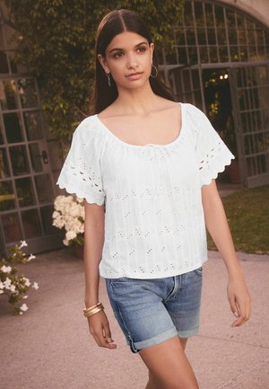 Witte katoenen top met oogletdetails, korte puffmouwen en een vierkante halslijn. Gecombineerd met denim shorts. Buitenscène met groene omgeving.