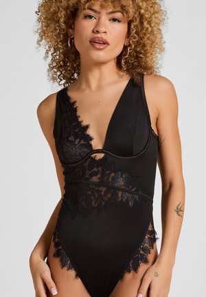 Frau mit lockigem Haar, die einen ärmellosen Body mit schwarzer Spitzenverzierung trägt und vor einem einfachen hellgrauen Hintergrund steht.