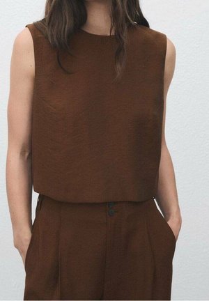 Kobieta ubrana w brązową bluzkę bez rękawów i pasujące, brązowe spodnie z wysokim stanem, ręce w kieszeniach na jasnym tle.