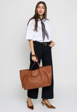 Mujer con blusa blanca, pantalones negros, pañuelo con estampado en el cuello y tacones marrones, sosteniendo un gran bolso marrón de cuero con doble asa.