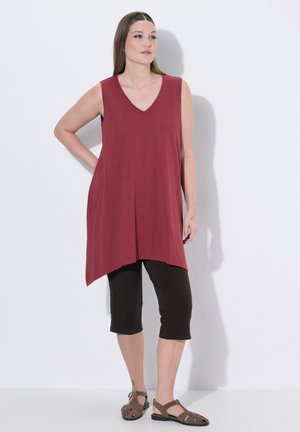 Femme debout portant une tunique bordeaux sans manches, des leggings noirs courts et des sandales marron fermées, sur fond blanc uni.