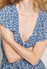 Blå blommig klänning med korta puffärmar, V-halsringning och knappar fram. Tyget har ett mönster av små vita blommor.