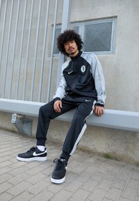 Μαύρο και γκρι σετ φόρμας Nike με φερμουάρ, λογότυπο και πλάγιες ρίγες; συνδυασμένο με μαύρα αθλητικά παπούτσια Nike με λευκές σόλες.