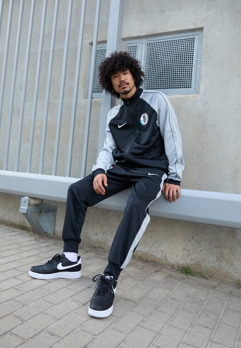 Μαύρο και γκρι σετ φόρμας Nike με φερμουάρ, λογότυπο και πλάγιες ρίγες; συνδυασμένο με μαύρα αθλητικά παπούτσια Nike με λευκές σόλες.