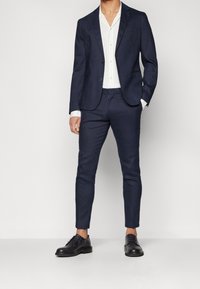 Námořnický modrý oblek s jedním knoflíkem, slim-fit kalhoty, bílá košile s vertikálními proužky a černé šněrovací boty.