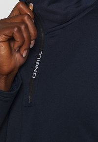 Mano di pelle scura che tira su la cerniera di una giacca O'Neill blu navy, con un focus sulla cerniera e sulla texture del tessuto.