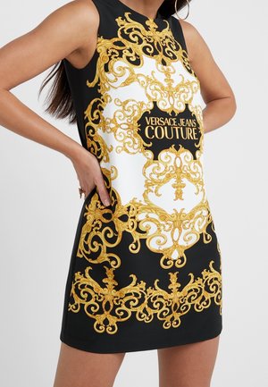 Czarna sukienka bez rękawów z białym środkiem i złotymi ozdobnymi barokowymi wzorami oraz tekstem "Versace Jeans Couture" z przodu.