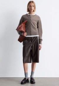 Brauner Strickcardigan mit Knöpfen, weißes T-Shirt, braune Leder-Shorts, graue Socken und Slipper. Hält eine geflochtene braune Tasche.