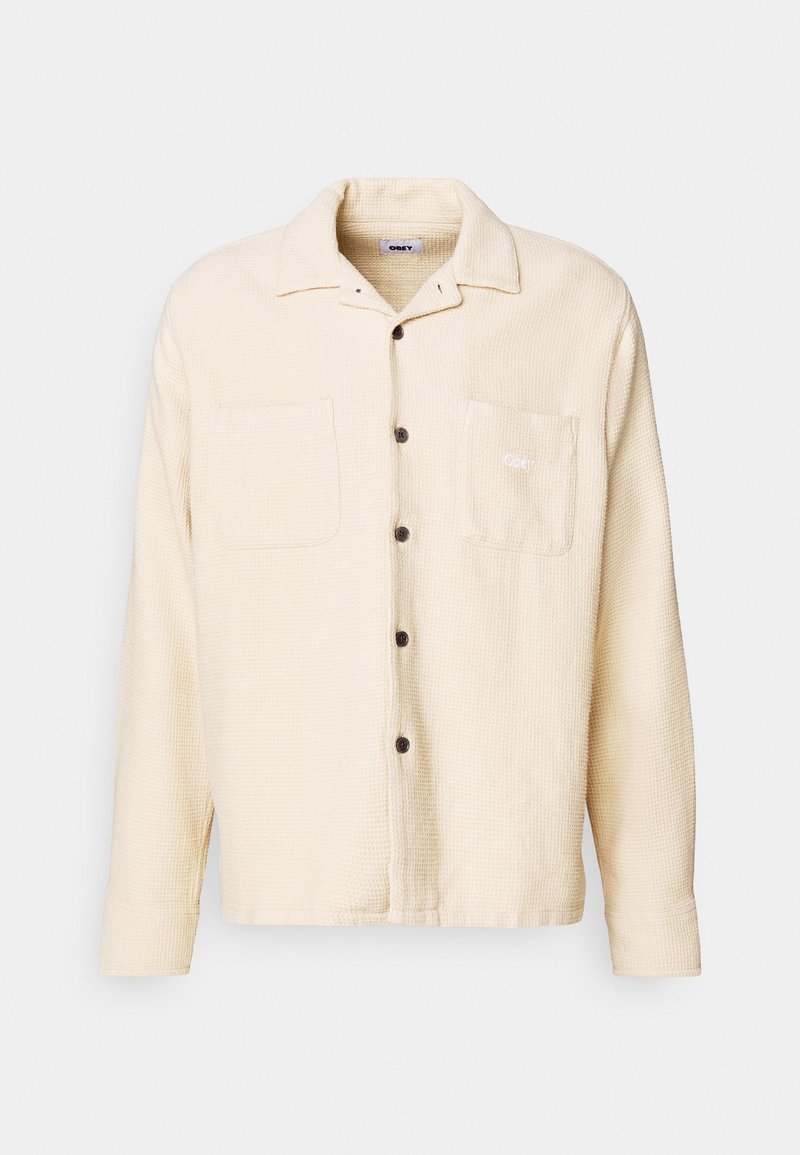 Ljust beige, texturerad skjortjacka med krage, fickor fram och mörka knappar. Har en broderad logotyp på vänster ficka.