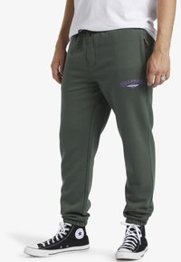 Groene sweatpants met een elastische tailleband, zijzakken en een paarse geborduurde logo. Geselecteerd met zwarte high-top sneakers.