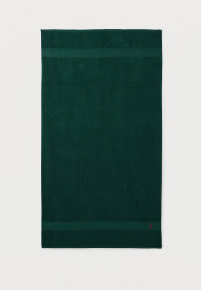 Σκούρα πράσινη ορθογώνια πετσέτα με δύο οριζόντιες λωρίδες κοντά στις άκρες και ένα μικρό κόκκινο λογότυπο στην κάτω δεξιά γωνία.