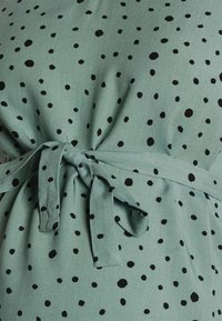 Tyrkysová látka s černými puntíky, zdobená zavázaným páskem v pase. Měkká textura s uvolněným střihem.