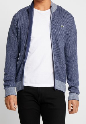 Mężczyzna noszący granatowy sweter rozpinany z teksturą, z jasnoszarymi mankietami i kołnierzem, na biały t-shirt i czarne spodnie.