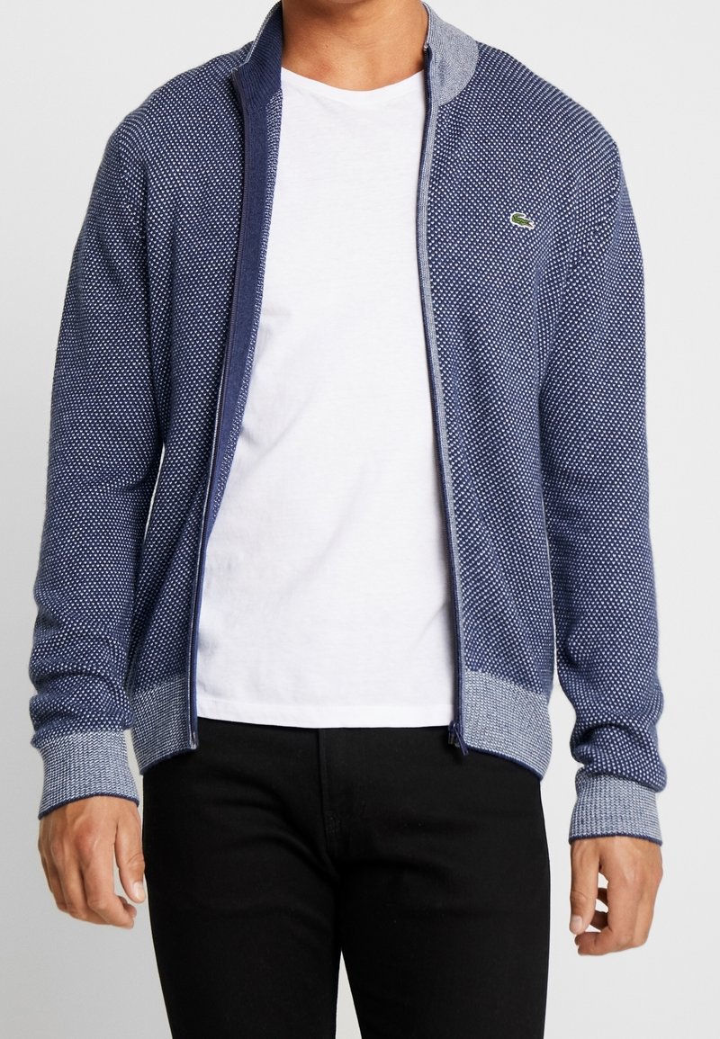 Homme portant un pull zippé texturé bleu marine avec des poignets et un col gris clair, par-dessus un t-shirt blanc et un pantalon noir.