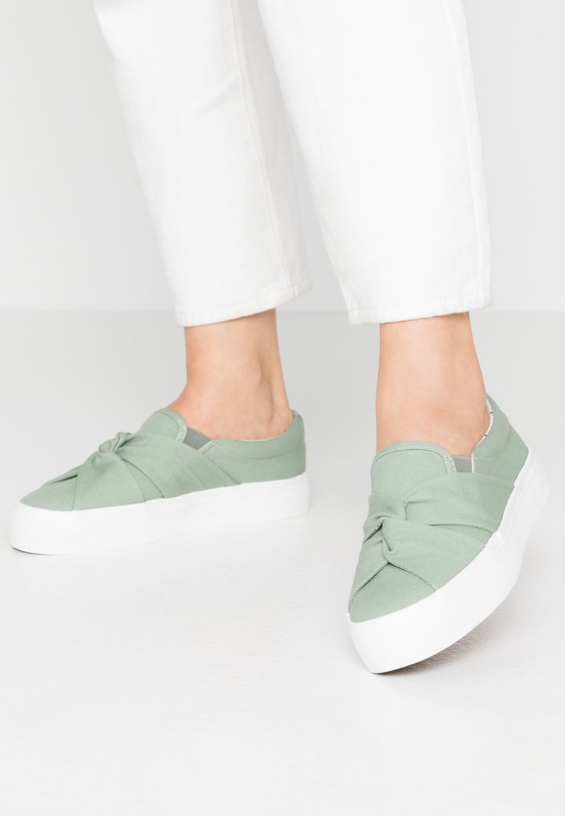 Personne portant un pantalon court blanc et des baskets légères vert clair à enfiler avec un détail de nœud et des semelles épaisses blanches sur un fond uni.