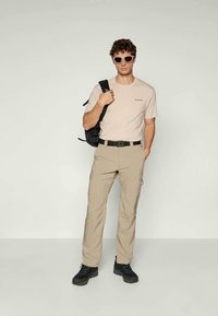 Světle béžové tričko s logem, pískové cargo kalhoty, černý opasek, černý batoh a tmavá obuv; outfit doplňují oversized sluneční brýle.