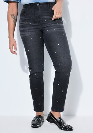 Persoon draagt zwarte spijkerbroek met kleine witte studs, gecombineerd met zwarte loafers met studs, staand tegen een lichte achtergrond.