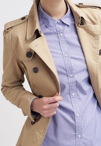 Person wearing a beige trench coat over a light blue button-up shirt, with one hand adjusting the coat near the chest.