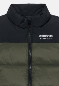 Nærbilde av en to-farget jakke med glidelås, der den øverste delen er svart og den nederste delen er olivengrønn, med "Outdoors Expedition" logo.