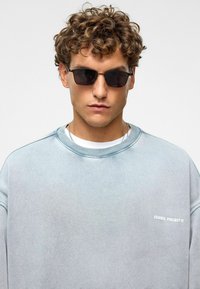 Lichtblauwe sweatshirt met ribbelkraag, met het logo "ESSENCE PROJECT" in het wit. Model draagt donkere zonnebril en een witte onderhemp.
