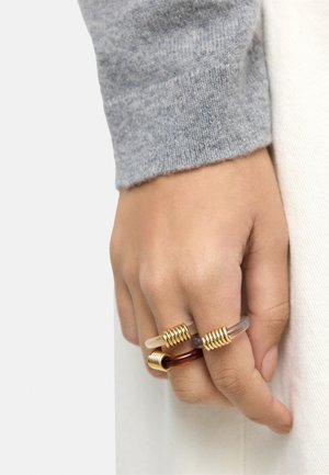 Ręka z dwoma przezroczystymi pierścieniami ozdobionymi złotymi spiralami na środkowym i serdecznym palcu, ubrana w szary długi rękaw i jasne spodnie.