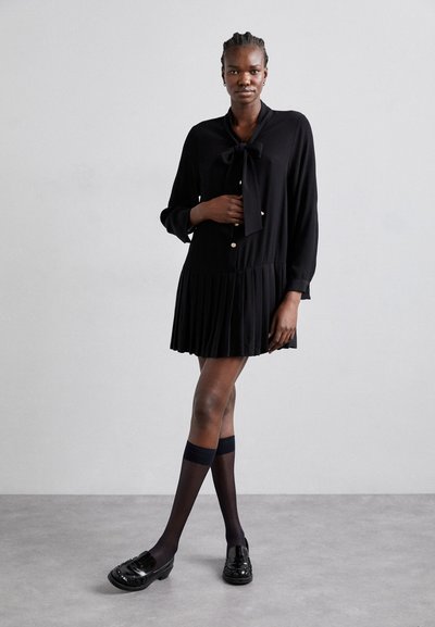 Robe plissée noire avec un col à nœud, manches longues, fermeture à boutons et assortie à des mocassins noirs et des chaussettes montantes. Texture de tissu lisse.
