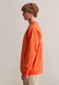 Pomarańczowa bluza z długimi rękawami, ściągaczami na mankietach i dolnym brzegu. Na przedzie znajduje się niewielki biały napis. Gładka faktura, casualowy krój.