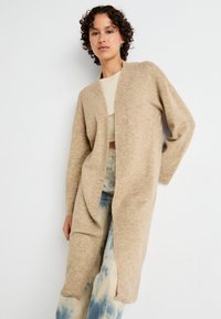 Beige lång cardigan i mjukt stickat material, med öppen front, långa ärmar och ribbade manschetter. Bärs över en ljus topp och mönstrade jeans.