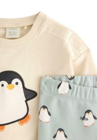 Cremfarbener Sweatshirt mit einem Pinguin-Druck und hellblauen Hosen, die ein Pinguin-Muster aufweisen. Weiches Baumwollmaterial, Etikett am Sweatshirt sichtbar.