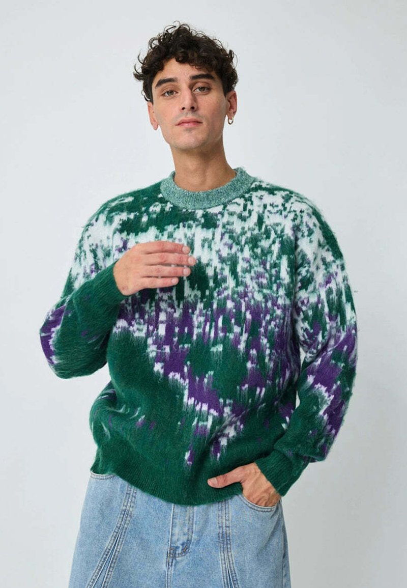 Pull vert tricoté avec un motif tie-dye en blanc et violet. Le design présente un col rond et des poignets côtelés. Texture douce.