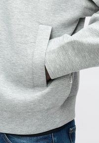 Ανοιχτό γκρι φούτερ με υφή, με πλαϊνή τσέπη και ριμπ μανσέτες. Συνδυάζεται με σκούρο τζιν.