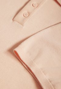 Polo shirt pêche clair avec un col, deux boutons et des manches courtes. Tissu doux et texturé, avec un détail en relief visible sur les bords.