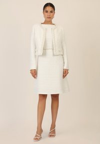 White tweed suit featuring a cropped jacket with pearl detailing and a flared skirt. Paired with white strappy heels for a complete look.