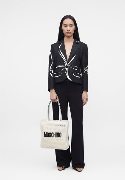 Femme portant un pantalon noir et un blazer noir avec des lignes abstraites blanches, tenant un sac cabas blanc Moschino avec un texte noir.