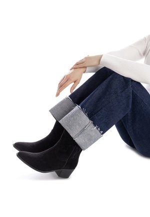Botines de ante negro con puntera puntiaguda y un tacón bajo y grueso, combinados con jeans de denim de pierna ancha que tienen un dobladillo deshilachado y vuelto hacia arriba.