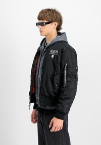Chaqueta bomber negra con diseño bordado "BERLÍN", bolsillos con cremallera y puños acanalados. Lleva puesta sobre una sudadera gris, combinada con pantalones oscuros.