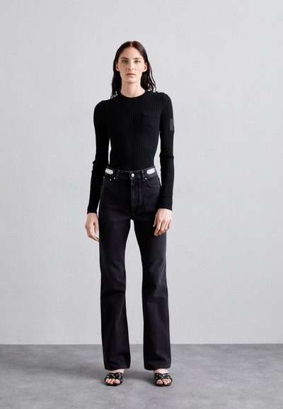 Black ribbed long-sleeve top with a pocket, paired with black straight-leg jeans featuring studded accents on the waistband. Black slide sandals.