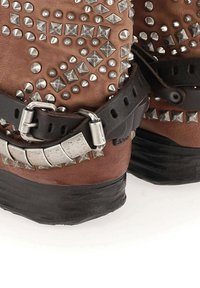 Bottes en cuir marron ornées de clous en argent, dotées d'une sangle noire avec une boucle argentée et de semelles en caoutchouc texturé.
