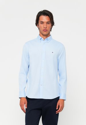 Light blue button-up shirt made of cotton. Features a traditional collar, long sleeves, and a small logo on the chest. Smooth texture.