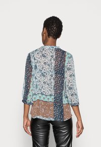 Blusa trasparente con un mix di motivi floreali e geometrici in verde teal, marrone e blu navy. Caratterizzata da maniche a 3/4 e scollo rotondo.