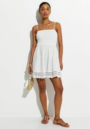 Mujer con vestido blanco sin mangas y mini con dobladillo de encaje, sandalias marrones, sosteniendo un bolso de paja, posando contra un fondo blanco liso.