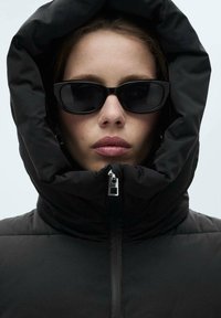 Geacă pufă neagră cu guler înalt și glugă, închidere cu fermoar și textură quilted. Asortată cu ochelari de soare subțiri negri.