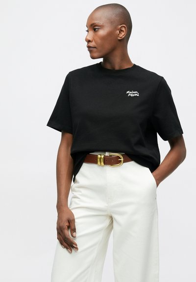 Osoba s krátkými vlasy, oblečená v černém tričku Maison Kitsuné, bílých kalhotách s vysokým pasem a hnědém opasku se zlatou sponou.