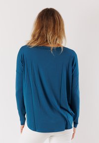 Maglietta a maniche lunghe realizzata in un tessuto liscio e elastico di un profondo colore teal. Presenta una vestibilità morbida e un collo rotondo, con un retro leggermente drappeggiato.