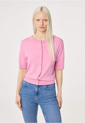 Vrouw met lang blond haar draagt een roze korte-mouwen vest met knopen en een blauwe spijkerbroek tegen een eenvoudige lichte achtergrond.