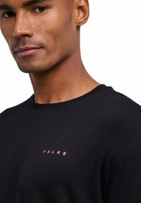 T-shirt noir à manches courtes avec un col rond, arborant un petit logo rose "FALKE" sur la poitrine gauche. Tissu doux et lisse.