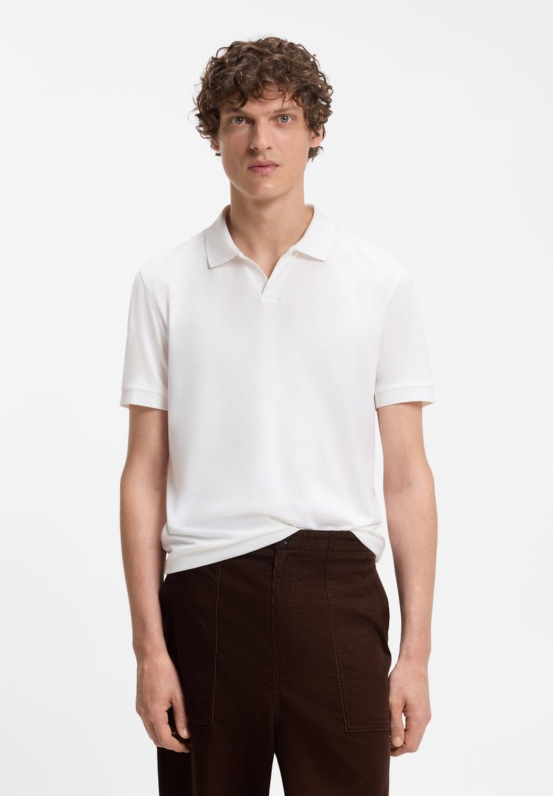 Homme aux cheveux bouclés portant un polo blanc à manches courtes rentré dans un pantalon marron foncé sur un fond blanc uni.