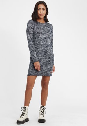 Robe en tricot à manches longues dans un motif chiné bleu marine et gris, avec un col rond et une fente sur le côté, associée à des bottes de combat blanches à lacets.