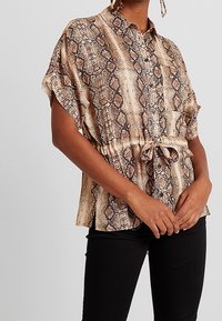 Camicia a maniche corte con bottoni, in un leggero pattern a stampa serpente marrone, con vita arricciata e un tessuto morbido e texturizzato. Abbinata a pantaloni neri.