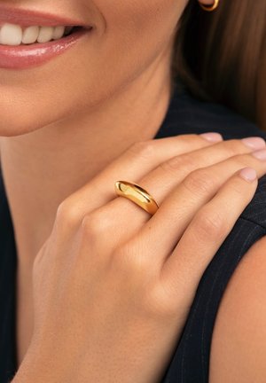 Anillo de color dorado con un diseño suave y redondeado, usado en una mano que descansa sobre una tela oscura, mostrando una superficie pulida y reflectante.